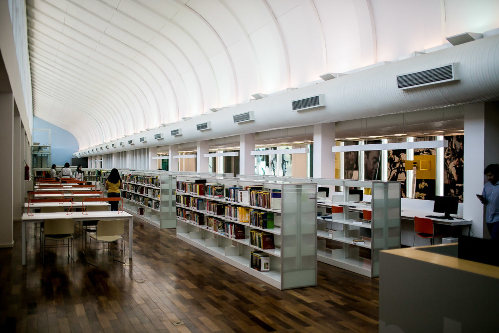 Interior moderno da Biblioteca Parque Estadual com teto curvo iluminado, estantes de livros, mesas de estudo e piso de madeira